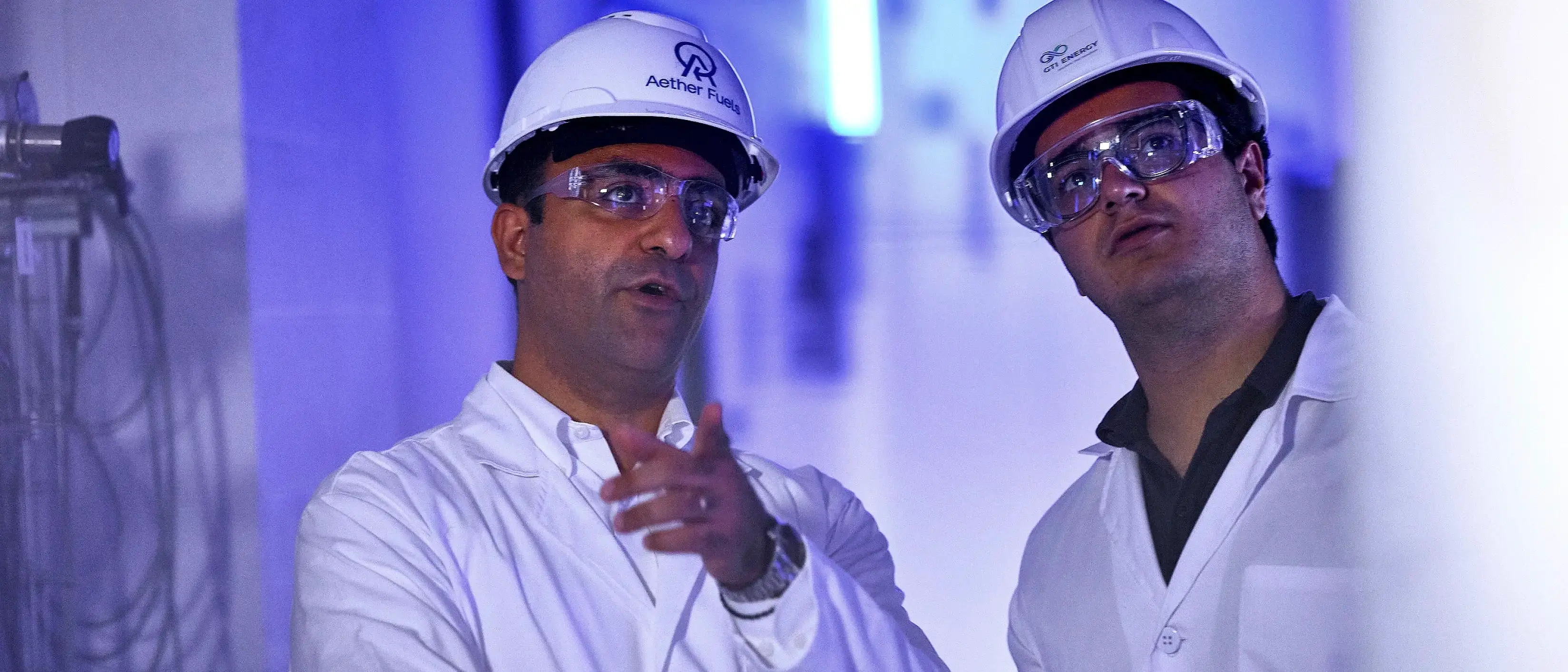 One man from Aether Fuels and another man from GTI Energy in hard hats and white coats focusing on something out of view.